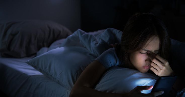 Young woman having sore and tired eyes when using smartphone while lying in bed at night